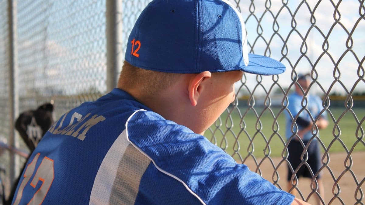 A young baseball player in uniform watches the game from behind a fence.