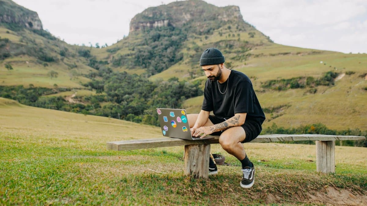 Young man with beard working on laptop outdoors in a scenic mountain setting. Ideal for remote work concepts.