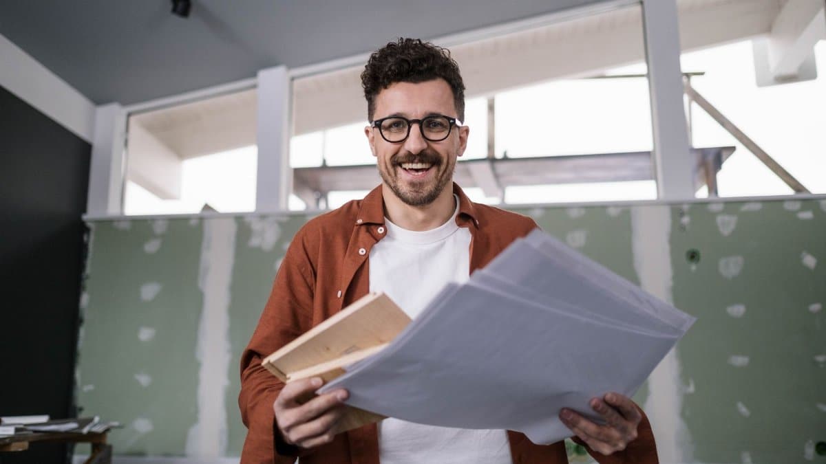 Smiling interior designer holding plans in a partially renovated room, conveying excitement and creativity.