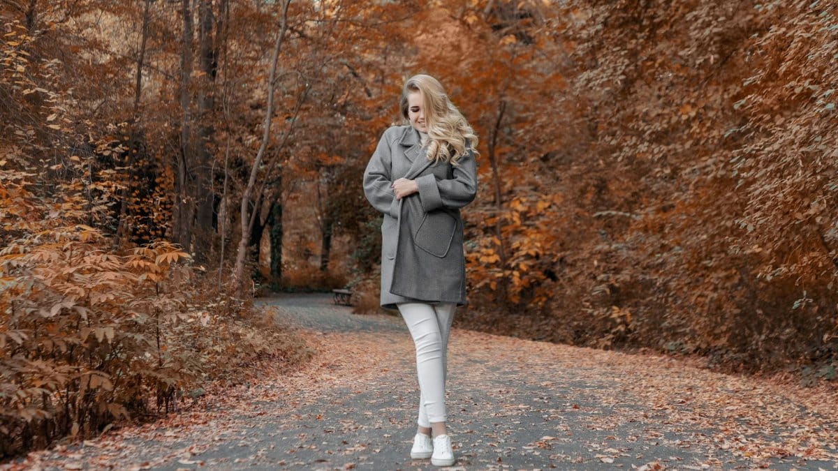 Full body happy young female bundling up in warm cozy coat while standing on asphalt road leading through autumn forest