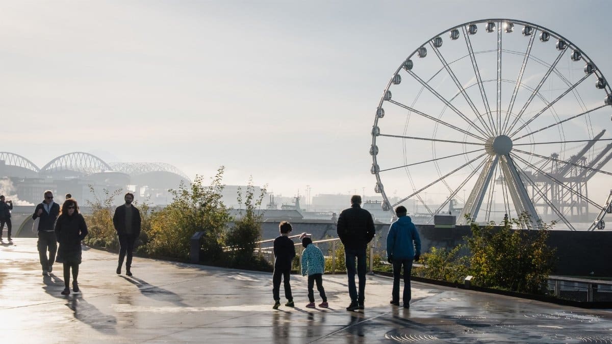 A group of people stroll along the waterfront with the Seattle Great Wheel in the background at dusk.
