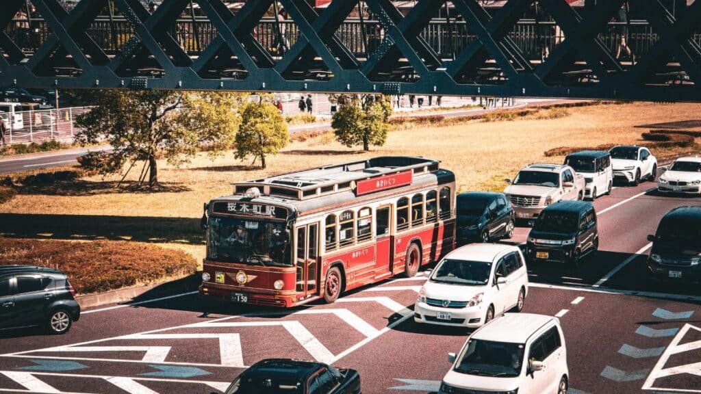 A vintage red bus travels on a busy street under a bridge, surrounded by cars.