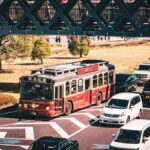 A vintage red bus travels on a busy street under a bridge, surrounded by cars.