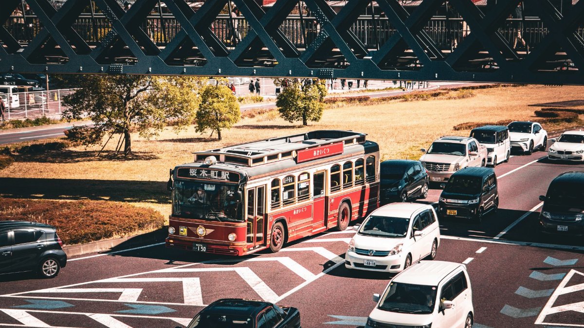 A vintage red bus travels on a busy street under a bridge, surrounded by cars.