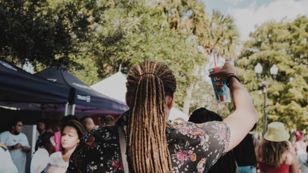 Back view of a man with dreadlocks holding a cup at an outdoor festival on a sunny day.