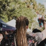 Back view of a man with dreadlocks holding a cup at an outdoor festival on a sunny day.