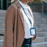 Side view of crop anonymous African American female entrepreneur with badge in classy brown coat standing near stairs with metal railings