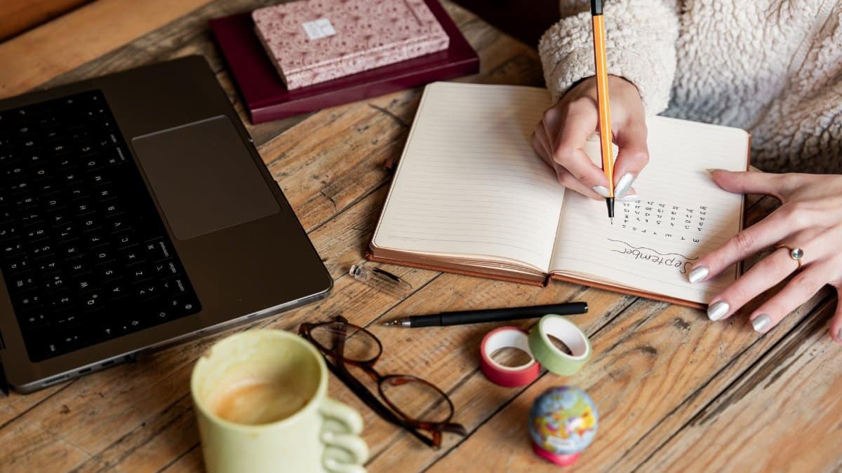 A woman planning her schedule in a notepad beside a laptop and coffee on a wooden desk.