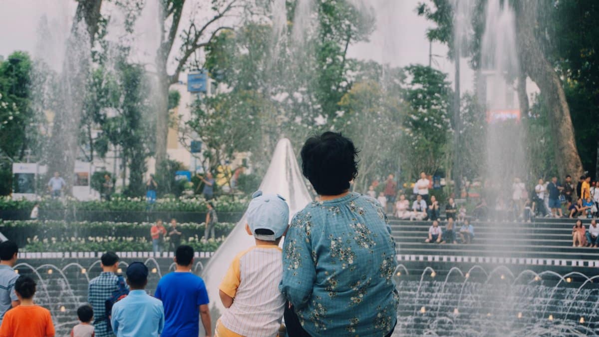 A family sits by a fountain in a bustling city park filled with visitors enjoying a sunny day.