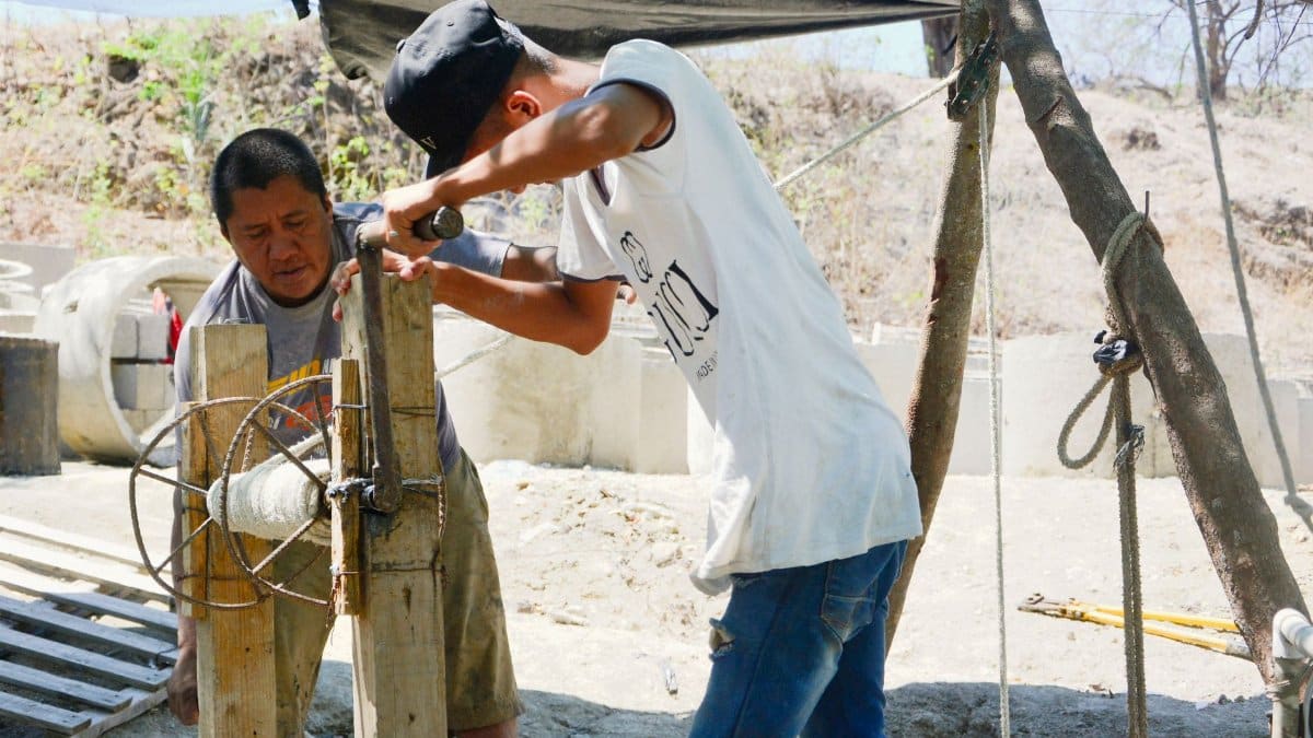 Two men working on a construction site, using a manual pulley under a tarp cover.