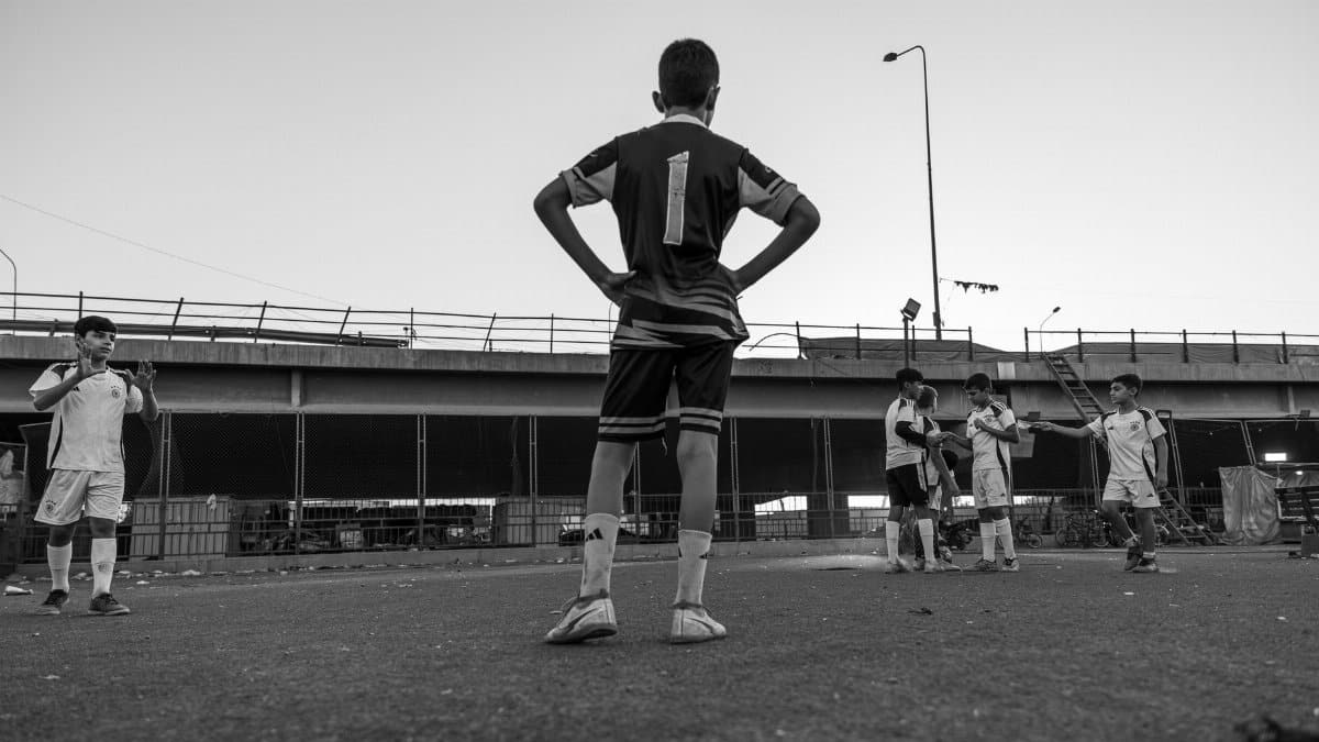 Black and white photo of boys playing soccer in an urban setting under a bridge.