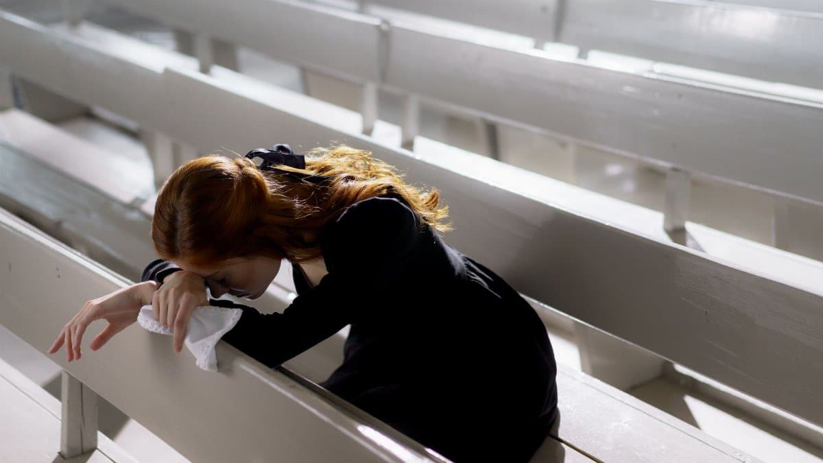 A woman in black clothes leans over pews, grieving alone in an empty church.