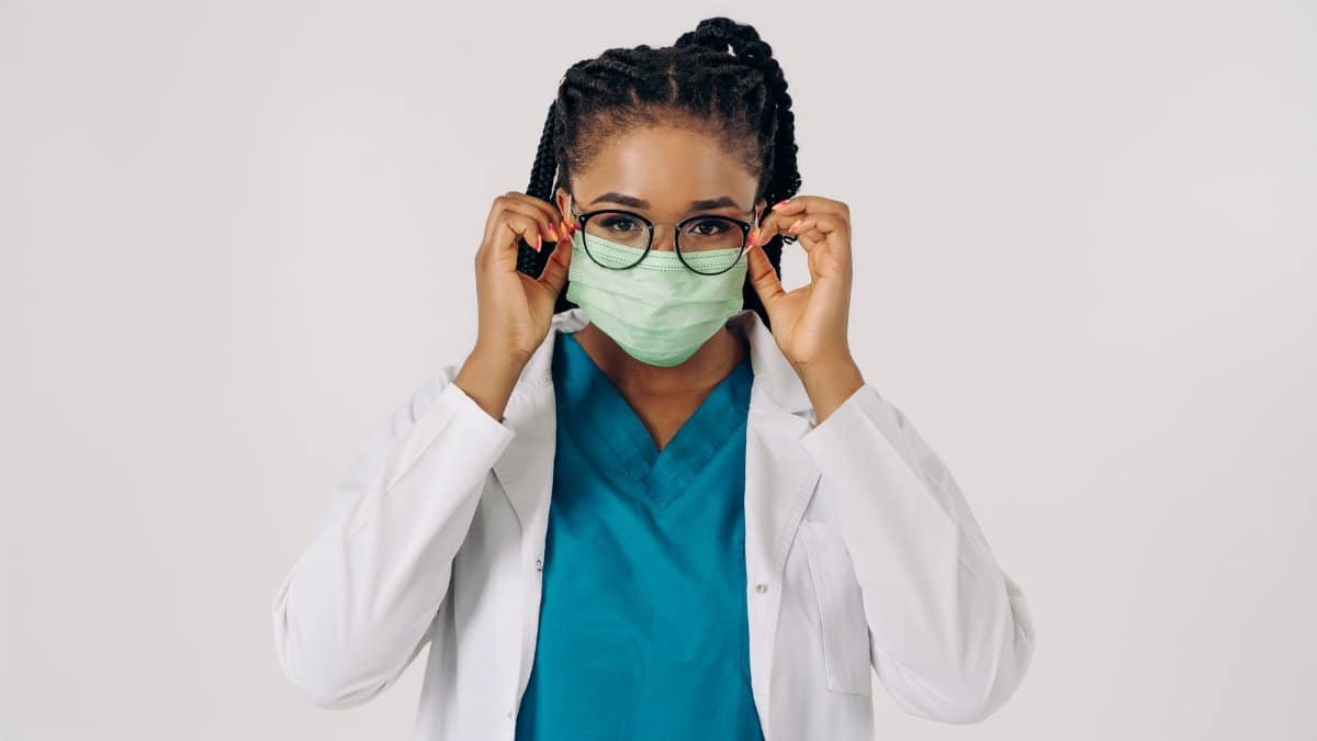 Healthcare professional in scrubs and mask adjusting glasses, studio setup.