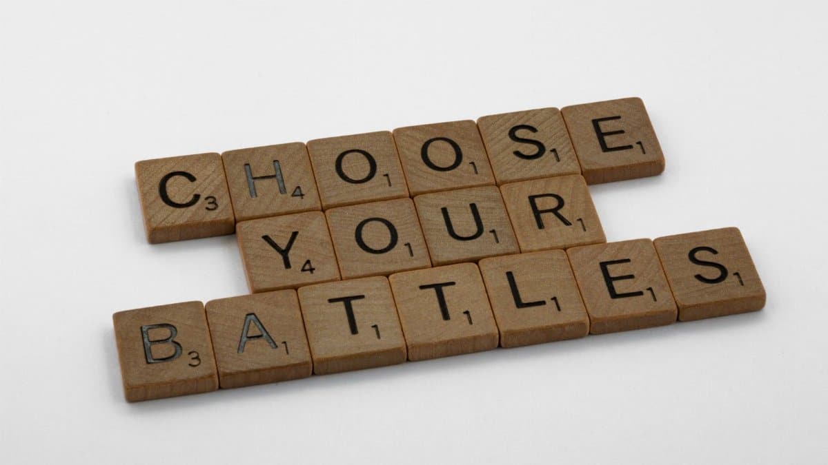 Wooden scrabble tiles spelling 'Choose Your Battles' on a white background.