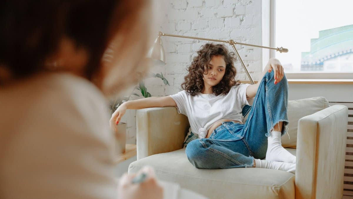 A young woman sits on a sofa during a therapy session, looking contemplative.
