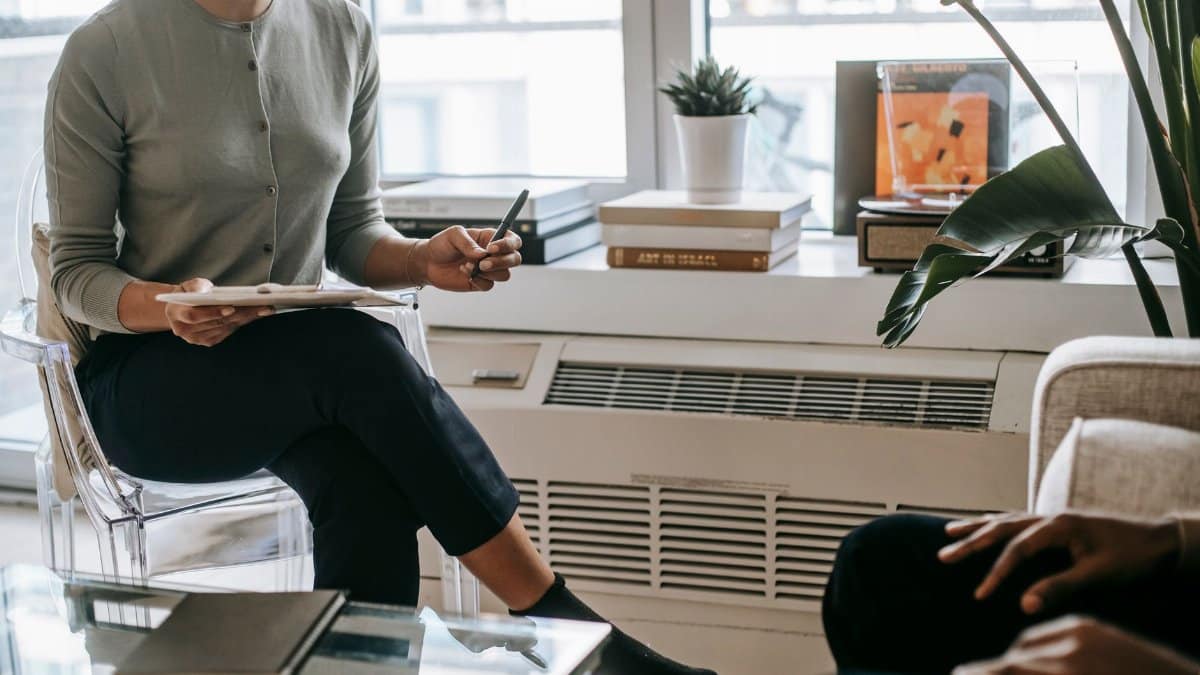 Unrecognizable female psychologist with crossed legs writing information on clipboard during appointment with anonymous ethnic patient in cozy light office