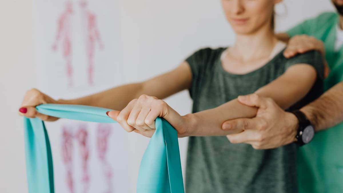 Crop anonymous orthopedist in wristwatch and uniform helping young woman in casual wear reaching arms with elastic tape in doctor office