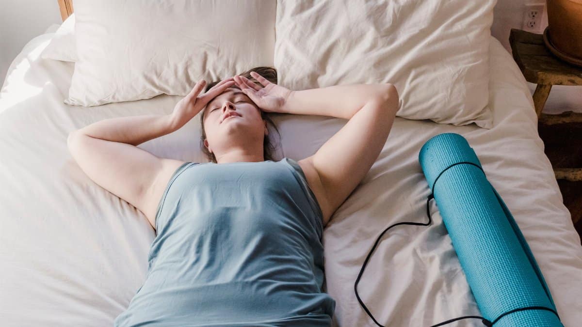 Woman lying on bed with a yoga mat beside her, appearing relaxed and comfortable indoors.