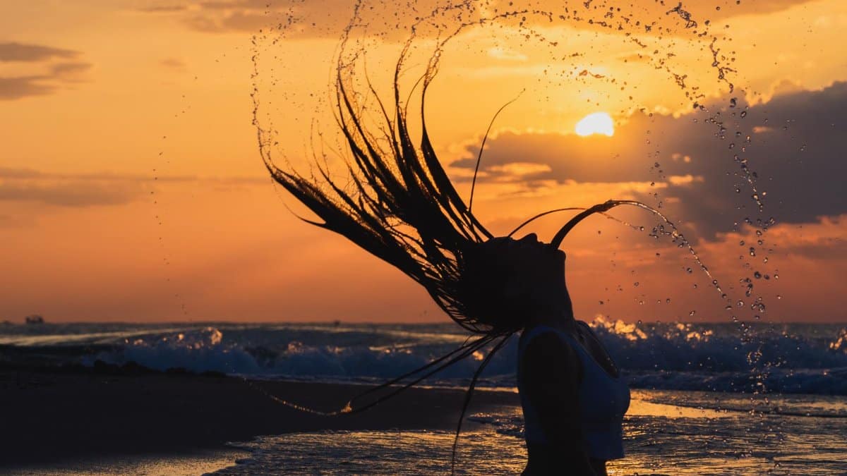 Silhouette of a woman flipping her hair at sunset, creating a water arc on the beach.