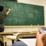 Student texting in a classroom while teacher is writing on the blackboard.