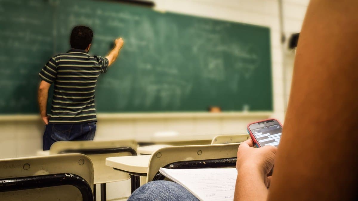 Student texting in a classroom while teacher is writing on the blackboard.