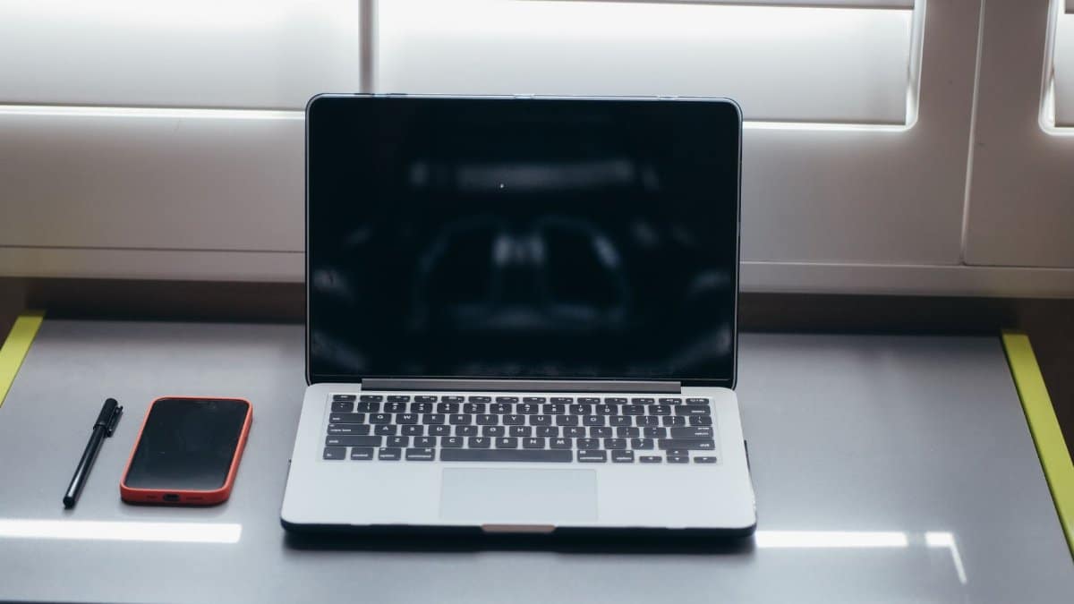 A minimalist desk setup featuring a laptop, smartphone, and pen under soft window light.