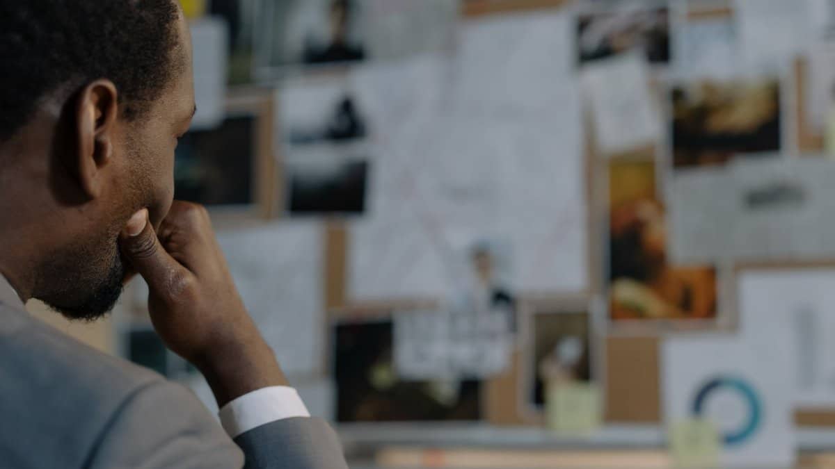 A detective in a suit examines an evidence board full of photos and documents.