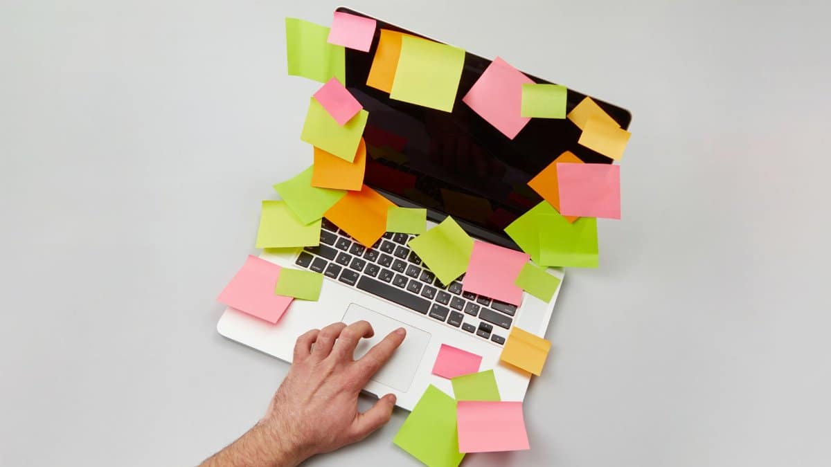 Hand on laptop surrounded by colorful sticky notes, depicting a busy and organized workspace.