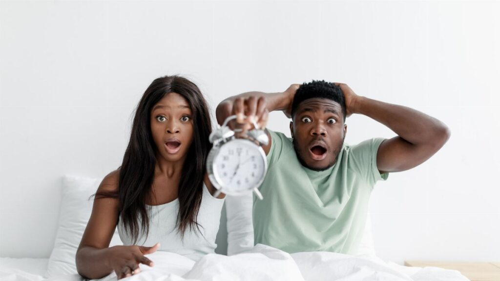 A surprised couple holding an alarm clock while sitting in bed, expressing shock.