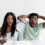 A surprised couple holding an alarm clock while sitting in bed, expressing shock.