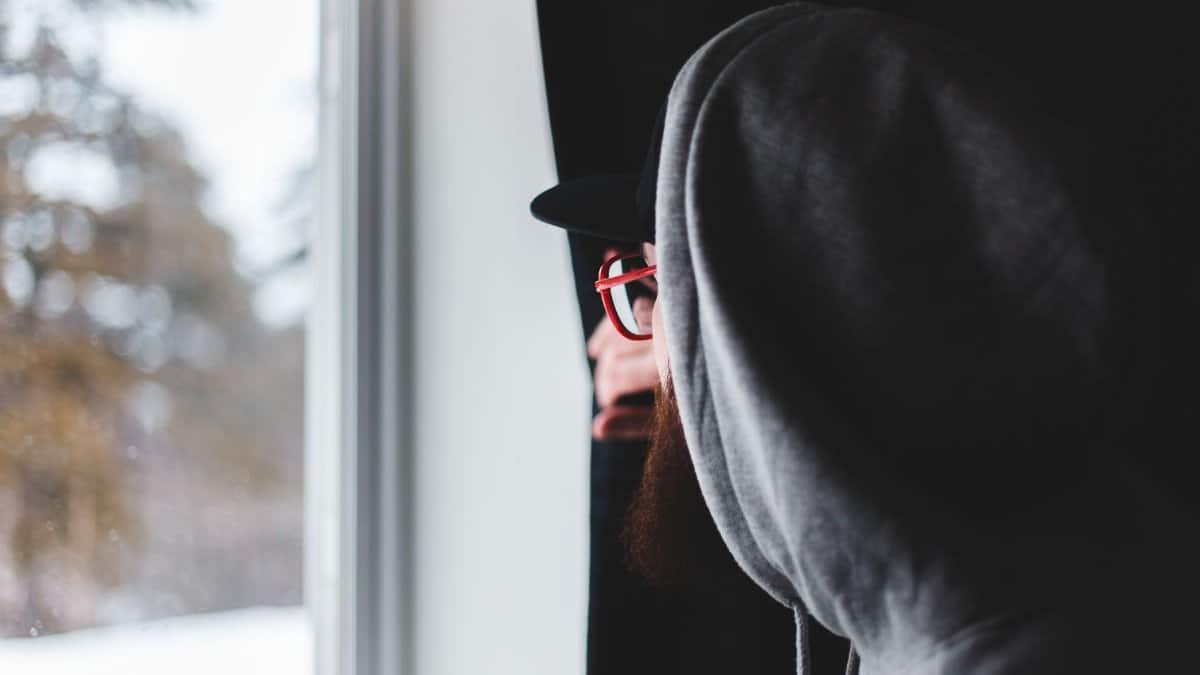 Side view of bearded man in glasses and hoodie pulling curtain and looking out window on winter day at home