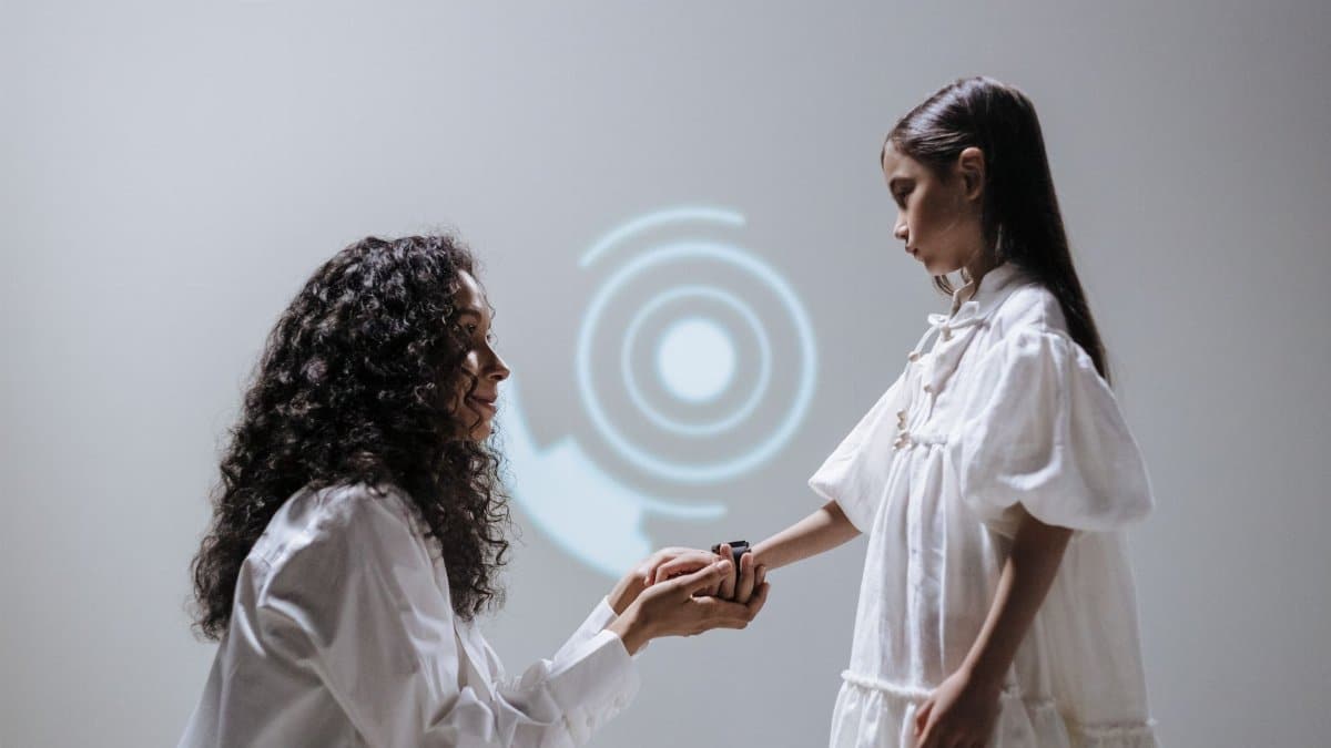 A mother kneeling and holding her daughter's hand, showcasing a smartwatch in a futuristic setting.