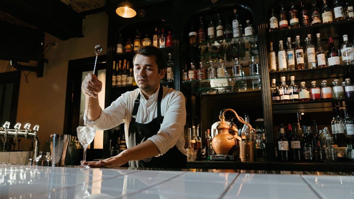 Male bartender mixing a cocktail behind a well-stocked bar. Atmospheric dim lighting indoors.