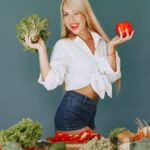 Cheerful woman in white shirt holding lettuce and pepper, celebrating fresh vegetables.