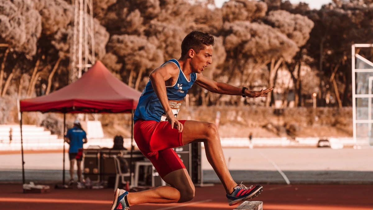 Dynamic shot of a young athlete in blue and red jumping a hurdle during a track race.