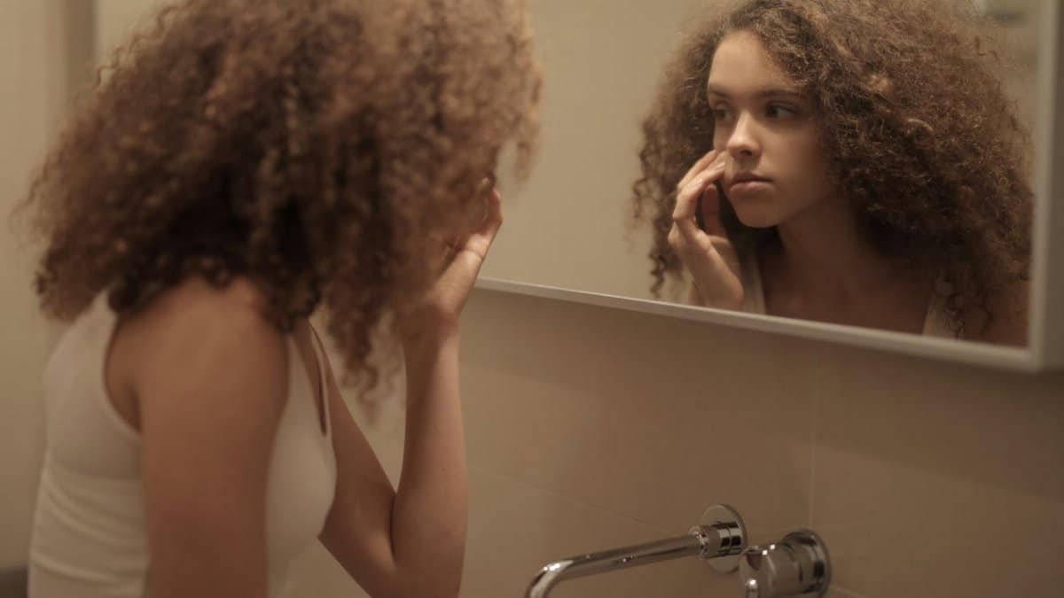 Teenage girl with curly hair examines her face in the bathroom mirror, reflecting contemplation.
