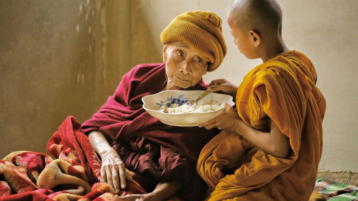A young monk offers food to an elderly person, highlighting compassion and care in Bangladesh.