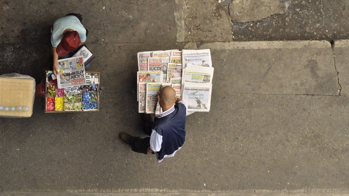 Street vendor arranging newspapers on a concrete sidewalk with candies and public interest articles.