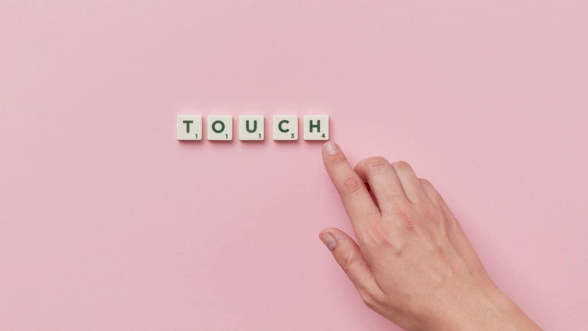 A hand pointing to Scrabble tiles forming the word 'TOUCH' on a pink surface.