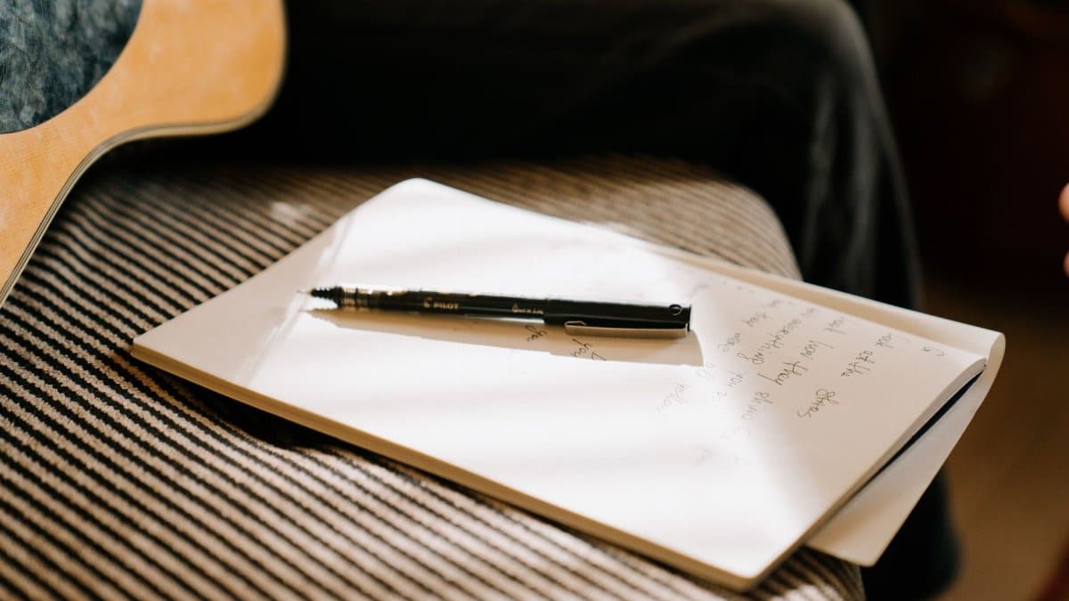 A close-up of a guitar, notebook, and pen suggests a moment of creative songwriting indoors.