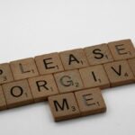 Wooden Scrabble tiles spelling 'Please Forgive Me' on a white surface, emphasizing forgiveness.