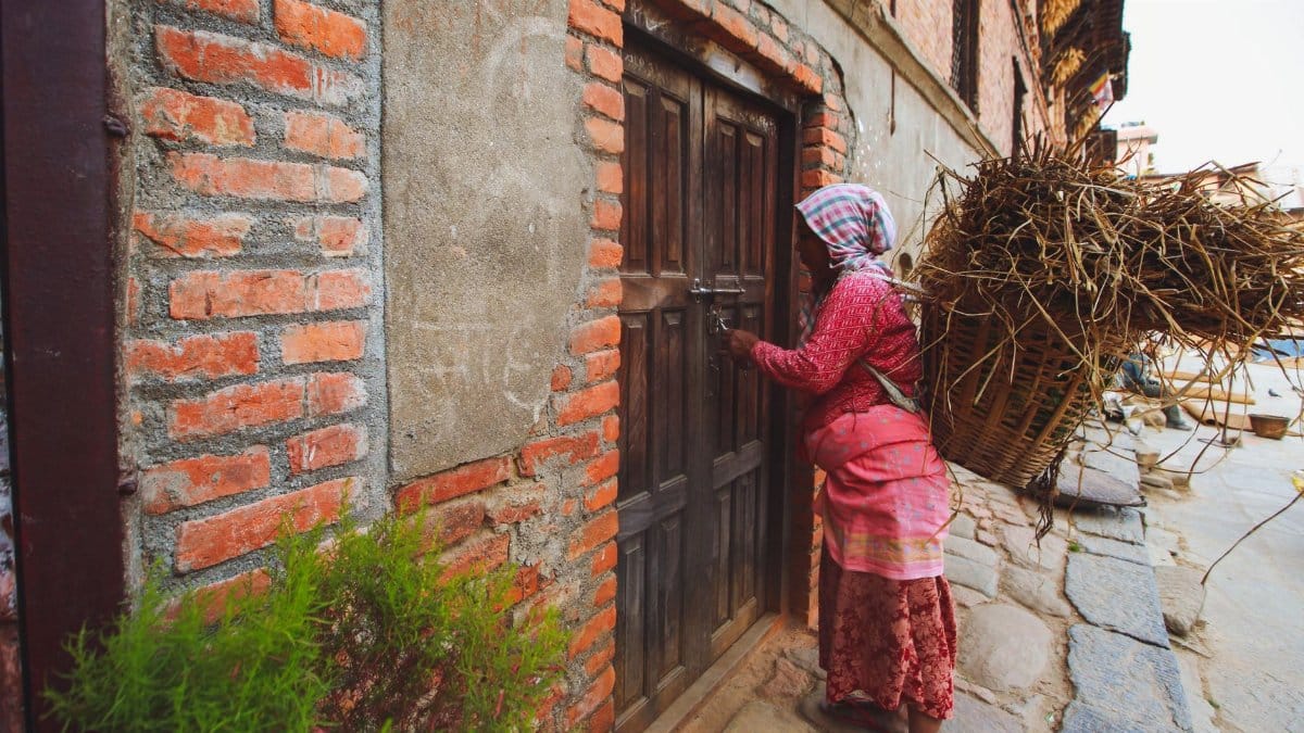 A woman carries a large basket of twigs on her back while opening a wooden door in a rustic village setting.
