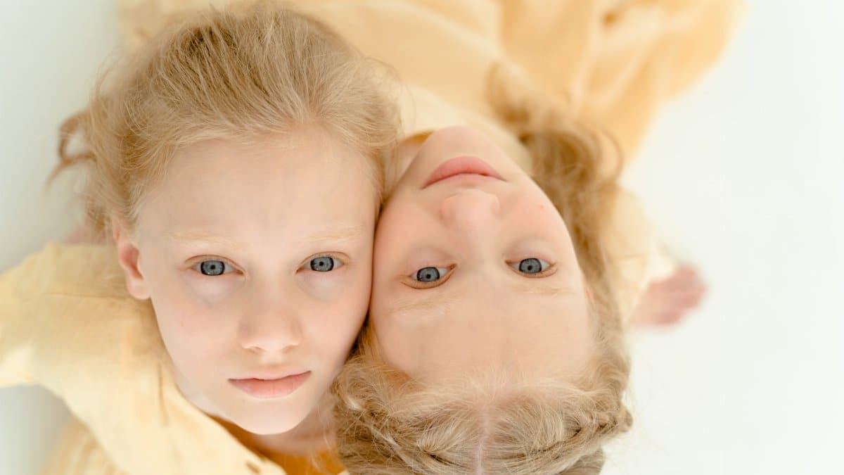 Charming portrait of twin girls with blond hair laying cheek to cheek in a light studio setting.
