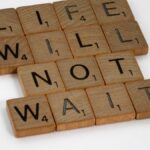 Wooden Scrabble tiles spelling 'Life Will Not Wait' on a white background.