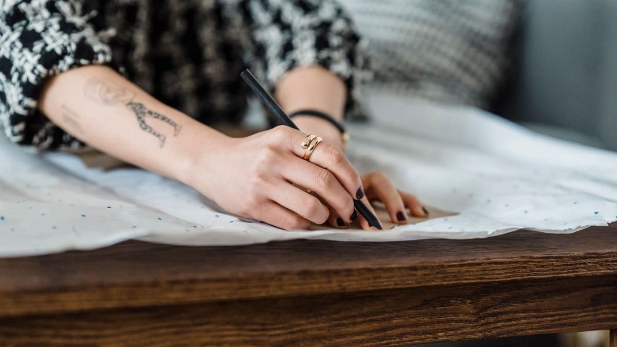 Crop unrecognizable female tailor with tattoo and pencil tracing on paper with carton pattern at table in house room