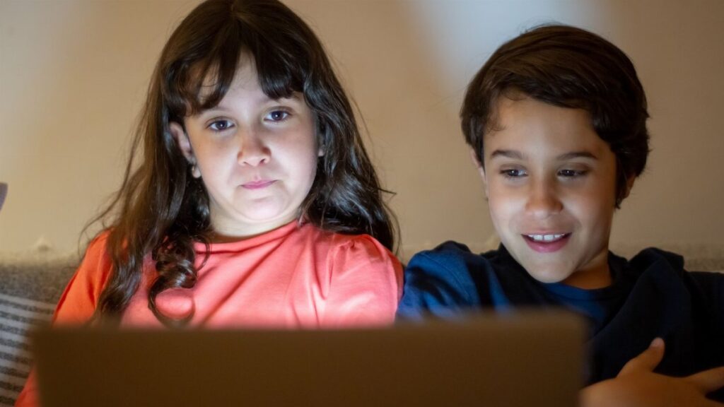 Two children watching a screen at home, focused and smiling, enjoying technology.