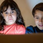 Two children watching a screen at home, focused and smiling, enjoying technology.