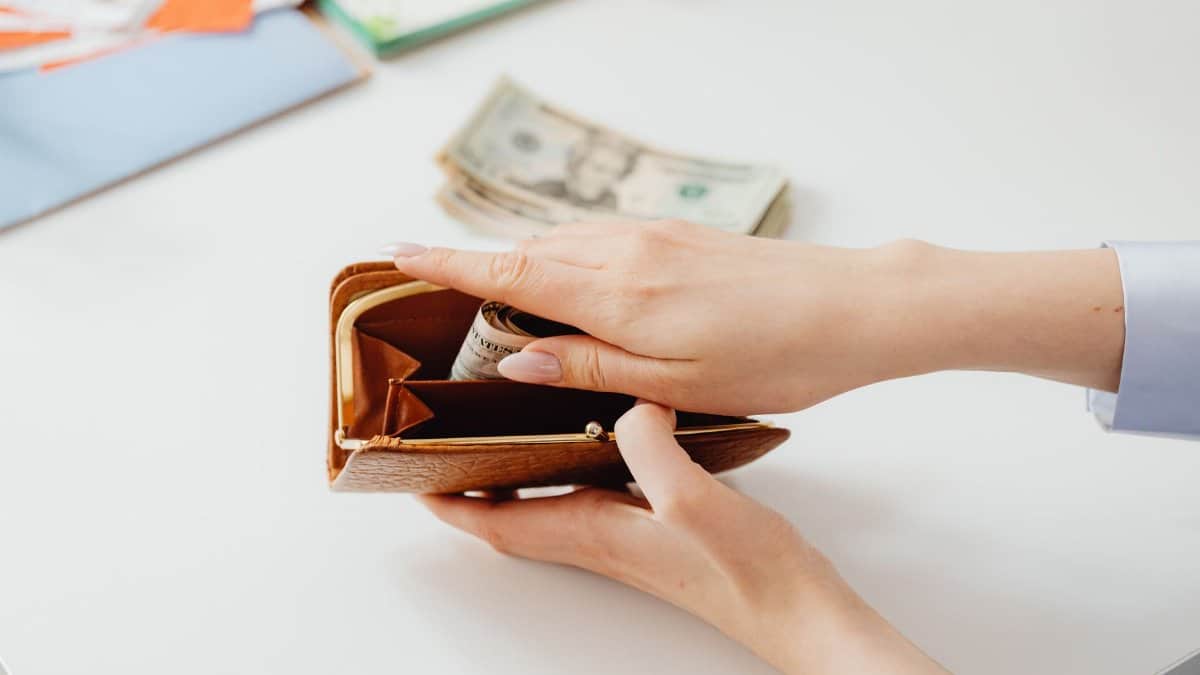 Close-up of hands placing cash into a leather wallet with bills on table.