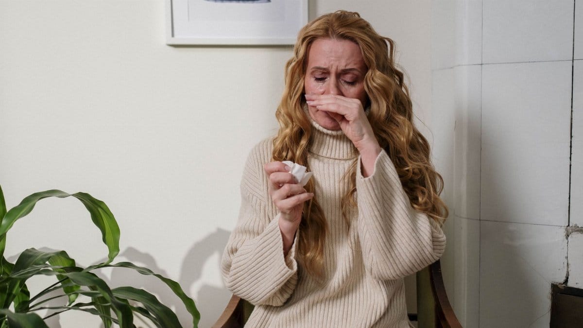 A woman with a runny nose sitting indoors, using a tissue to relieve her allergies.