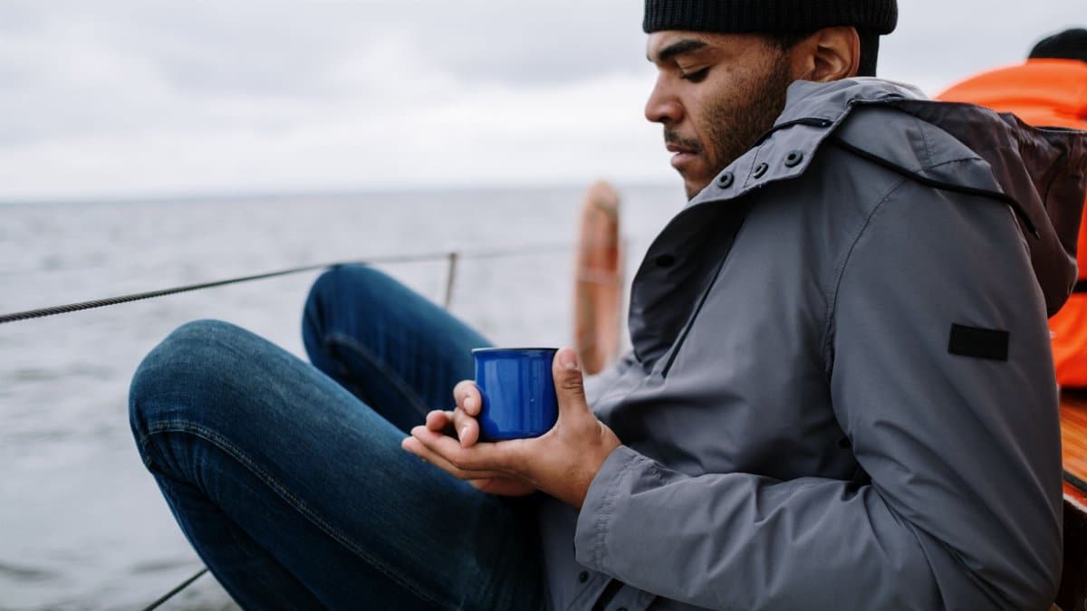 A man in a jacket relaxes on a sailboat holding a hot drink in a blue cup.